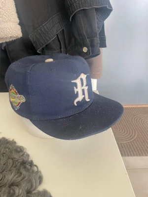 Mörkblå keps med broderad R-logga - Snygg mörkblå keps med vit broderad R-logga framtill och färgglad patch på sidan. Klassisk flat brim och justerbar baksida. Perfekt accessoar för en streetwear-look. New Era keps.