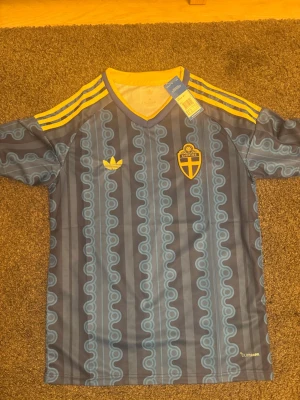 Sverige retro fotbollströja Adidas - Retroinspirerad fotbollströja från Adidas med Sveriges landslagsemblem på bröstet. Tröjan är blå med gula detaljer, har klassiska Adidas-ränder på axlarna och ett unikt mönster i ljusblått. Tillverkad i lätt och ventilerande material, perfekt för träning eller match.