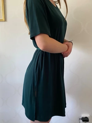 Mörkgrön kortklänning från Lindex - En enkel och stilren mörkgrön klänning från Lindex med korta ärmar och rund halsringning. Klänningen har slitsar på båda sidor och är gjord i ett mjukt, stretchigt material som känns skönt mot huden. Perfekt för en avslappnad look. Bandet har en ljusrae nyans av grön. Klänningen är i storlek L men passar lika bra en person som vanligt vis har storlek S. Plagget säljs även på en annan säljsida.