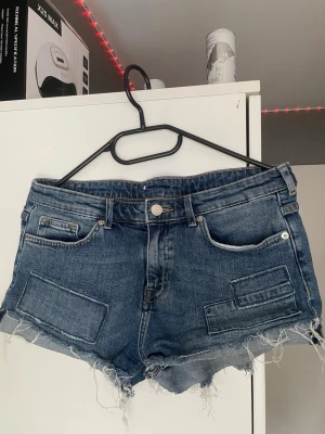 Blå jeansshorts med fransig kant - Snygga blå jeansshorts med rå, fransig kant och slitna detaljer. Shortsen har klassiska fickor fram och bak samt extra dekorativa fickor framtill. Perfekta för en avslappnad och trendig look. Vet inte vart den är ifrån då jag köpte den på secondhand och det har ingen lapp på sig så vet ej vilken storlek men skulle nog säga 36/34