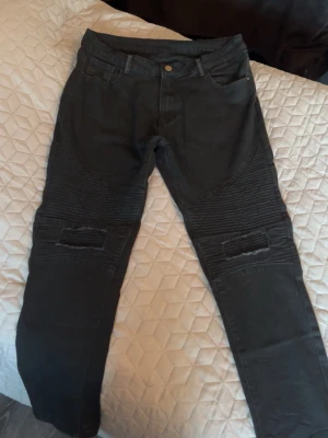 Svarta jeans med detaljer - Säljer ett par svarta jeans med ribbade partier över knäna och förstärkta detaljer. Jeansen har dragkedjegylf, fem fickor och smal passform. Perfekta för en edgy streetstil och funkar till sneakers, storlek w34 l33