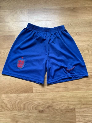 Blå England Nike fotbollsshorts - Snygga blå fotbollsshorts från Nike med England-emblem och röd Nike-logga på framsidan. Shortsen har elastisk midja och är tillverkade i ett lätt och bekvämt material som passar perfekt till träning eller match.