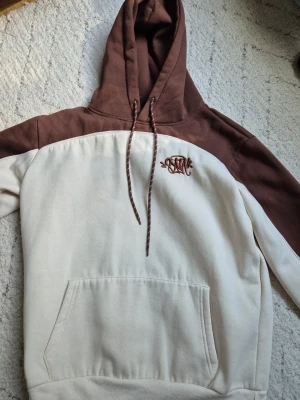 Brun och beige hoodie  - Snygg hoodie i tvåfärgad design med brun huva och axelparti samt beige kropp och ärmar. Broderad detalj på bröstet och klassisk känguruficka framtill. Mjuk bomullsblandning och dragsko i huvan för extra stilpoäng.