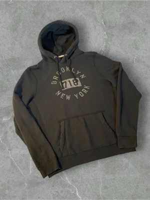 Svart hoodie Brooklyn New York |  XL - Svart hoodie med tryck 'Brooklyn 718 New York' på bröstet. Klassisk känguruficka framtill och justerbar huva med snören. Tillverkad i mjukt bomullsmaterial, perfekt för en avslappnad streetwear-look.