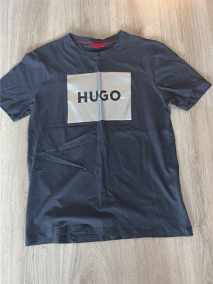 Mörkblå t-shirt från Hugo Boss - Cool svart t-shirt från Hugo med stor vit logga på bröstet. Klassisk rund hals och korta ärmar. Perfekt för dig som gillar stilrena plagg med tydlig branding. Materialet är mjukt och känns skönt mot huden. Nästan aldrig använd så nyskick 10/10!