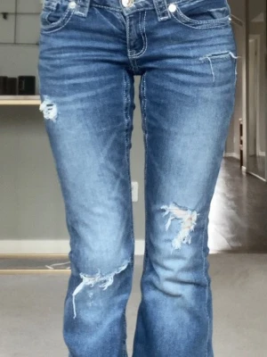 Blå bootcut jeans med slitningar - Snygga blå bootcut jeans med slitna detaljer och hål vid knäna. Jeansen har klassisk femficksdesign, låg midja och markerade sömmar. Perfekta för en avslappnad och trendig look.