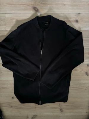 Svart cardiganjacka från Massimo Dutti - Stilren svart cardiganjacka från Massimo Dutti med dragkedja framtill och ribbad krage. Jackan är tillverkad i mjukt material och har en minimalistisk design som passar perfekt till olika outfits.