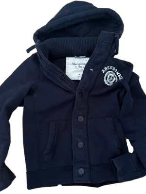 Mörkblå hoodie från Abercrombie & Fitch - Snygg mörkblå hoodie från Abercrombie & Fitch med knäppning framtill och broderad logga på bröstet. Hoodien har en stor huva, ribbade muddar och två fickor fram. Tillverkad i mjukt bomullsmaterial som känns skönt mot huden.