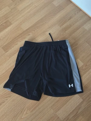 Svarta träningsshorts från Under Armour - Säljer ett par svarta träningsshorts från Under Armour med grå sidopanel och vit logga. Shortsen har elastisk midja med snörning och är tillverkade i ett lätt, syntetiskt material som andas. Perfekta för gymmet eller löprundan.