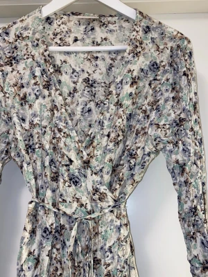 Blommig omlottklänning med knytband - Supersöt blommig omlottklänning i ljusa toner med inslag av blått, grönt och beige. Klänningen har lång ärm, v-ringning och knytband i midjan för en smickrande passform. Perfekt för vår och sommar! 