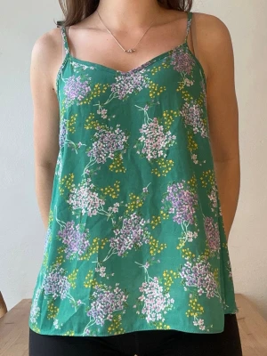 Grön blommig linnetopp - Snygg grön linnetopp med smala axelband och blommigt mönster i lila, rosa och gult. Toppen har en lös passform och v-ringad halsringning, perfekt för varma dagar.