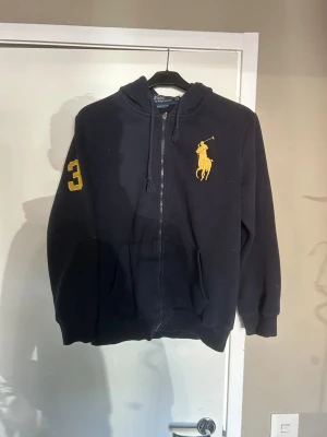 Marinblå hoodie från Polo Ralph Lauren - Snygg marinblå hoodie från Polo Ralph Lauren med dragkedja, stor gul logga på bröstet och siffran 3 på ärmen. Klassisk custom fit-modell med huva och ficka. Perfekt för en avslappnad och sportig stil.