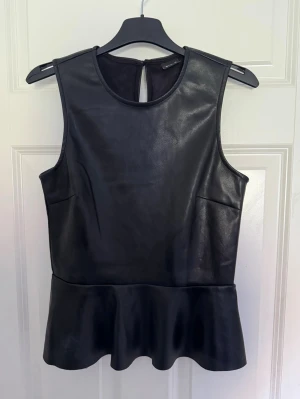 Svart ärmlös peplumtop i skinnimitation - Snygg svart ärmlös peplumtop i skinnimitation med rund halsringning och öppning bak i nacken. Perfekt för att lyfta en outfit med en edgy touch. Sitter figurnära upptill och har en volang nedtill.