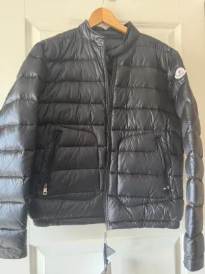 Svart dunjacka från Moncler - Snygg svart dunjacka från Moncler med quiltad design och hög krage. Jackan har två dragkedjeförsedda fickor framtill och Moncler-logga på vänster ärm. Perfekt för kalla vinterdagar och riktigt stilren look. Passar S/ liten M
