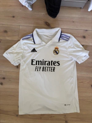 Real Madrid matchtröja Adidas vit - Säljer en vit Real Madrid matchtröja från Adidas med klubbmärke på bröstet och svarta detaljer. Tröjan har korta ärmar, krage och lila Adidas-ränder på axlarna. Materialet är lätt och andas, perfekt för fotboll eller träning.