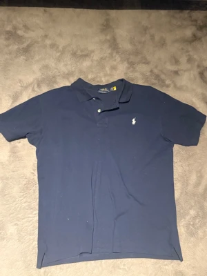 Marinblå pikétröja från Polo Ralph Lauren - Klassisk marinblå pikétröja från Polo Ralph Lauren med korta ärmar och vit broderad logga på bröstet. Tröjan har krage och två knappar framtill. Perfekt för en stilren och avslappnad look.