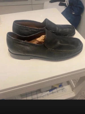 Svarta loafers i läder - Klassiska svarta loafers i äkta läder med stilren design och rundad tå. Skorna har en låg klack och slät sula, perfekta för en clean och tidlös look. Passar till både jeans och chinos för en avslappnad men ändå uppklädd stil.