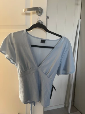 Ljusblå omlottopp från Ginatricot - Säljer en ljusblå omlottopp från Ginatricot med v-ringning och korta ärmar. Toppen har knytband i midjan som ger en snygg passform och är tillverkad i mjuk bomull. Använd 1 gång så den är som ny!