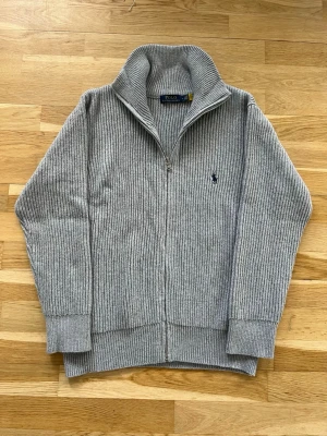 Grå ribbstickad zip-tröja Polo Ralph Lauren - Ribbstickad grå zip-tröja från Polo Ralph Lauren med hög krage och broderad logotyp på bröstet. Tröjan har långa ärmar, breda muddar och är tillverkad i mjukt stickat material. Perfekt för lager-på-lager och snygg till jeans.