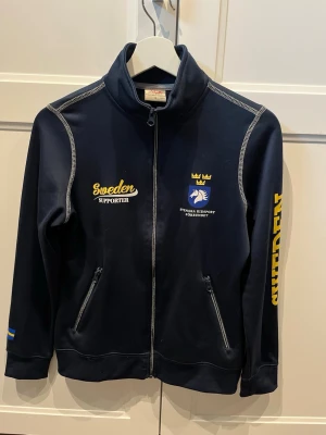 Svenska ridsportförbundet tröja - Mörkblå ziptröja med gul text 'Sweden Supporter' och Svenska Ridsportförbundets logga på bröstet. Dragkedja framtill, två fickor med dragkedja och tryck på ena ärmen. 