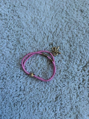 Rosa armband med rosettberlock - Sött armband med rosa snöre och guldfärgade detaljer. Armbandet har en dekorativ rosettberlock i metall och små guldfärgade pärlor. Perfekt accessoar för att ge din outfit en lekfull touch.