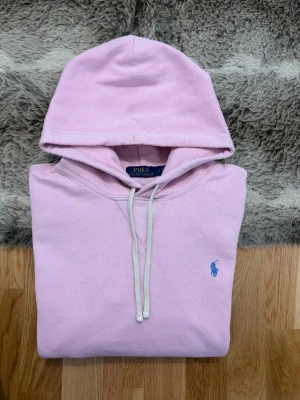 Ljusrosa Polo Ralph Lauren Hoodie - Storlek L - Hej! 👋 Säljer en fin ljusrosa Polo Ralph Lauren-hoodie i storlek L. 🌸 En härlig färg som verkligen piggar upp garderoben! Tröjan är i gott skick men har spår av användning: det finns några små prickar/fläckar samt ett professionellt lagat hål (se bilder). Det är inget som stör helhetsintrycket, men priset är satt därefter. ✨ 100% äkta med både tvättlappar och QR-kod för verifiering ✅ Perfekt som en skön och billig vardagströja! Fraktar snabbt! 🚛📦💬💯 
