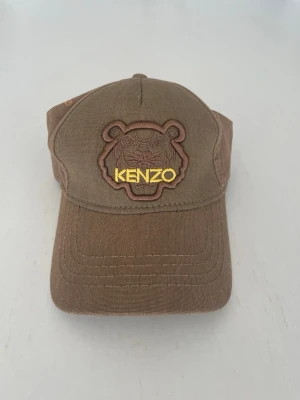 Brun Kenzo keps med tigerbrodyr - Snygg brun keps från Kenzo med broderad tiger och gul logotyp framtill. Justerbar rem bak för perfekt passform. Fick kepsen av en polare för ett tag sedan men har inte använt den då den ej passade mig