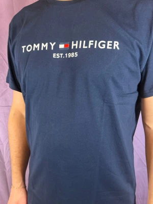 Blå Tommy Hilfiger t-shirt - Snygg marinblå t-shirt från Tommy Hilfiger med klassiskt logotryck och texten 'EST.1985' på bröstet. T-shirten har rund hals och korta ärmar, perfekt för en avslappnad och stilren look. Materialet är mjukt och skönt bomull.