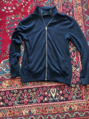 Mörkblå ziptröja från Jack & Jones - Mörkblå ziptröja från Jack & Jones med hög krage och hel dragkedja. Tröjan har ribbade muddar vid ärmslut och nederkant samt två fickor framtill. Perfekt för lager-på-lager och enkel att matcha med jeans eller chinos.