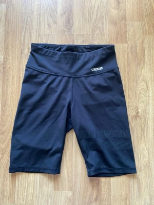 Svarta shorts från Stronger - Svarta shorts från Stronger med hög midja och diskret logga framtill. Tillverkade i stretchigt och följsamt material som passar perfekt för träning eller sportiga aktiviteter. Snygg och enkel design med bekväm passform.
