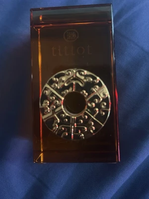 Tittot parfymflaska med dekor - Unik parfymflaska från Tittot i mörk bärnstensfärgat glas med en rund, silverfärgad dekor framtill. Flaskan har en rektangulär form och dekoren har ett asiatiskt inspirerat mönster. Perfekt för dig som gillar snygga och annorlunda parfymflaskor.