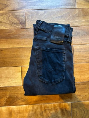Svarta Replay anbass - Säljer ett par svarta slim fit jeans från Replay med klassisk femficksdesign och läderpatch bak i midjan. Jeansen har smal passform och är tillverkade i stretchigt denimtyg för extra komfort. Perfekta för en trendig och avslappnad stil.