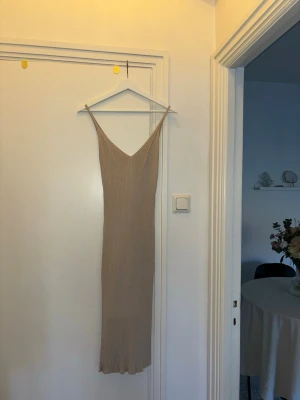 Beige ribbad långklänning - Säljer en beige ribbad långklänning med smala axelband och v-ringning. Klänningen har en figurnära passform och är tillverkad i ett stretchigt material som ger en snygg siluett. Perfekt för dig som gillar minimalistisk och stilren design.