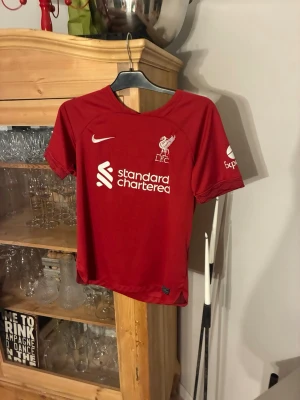 Liverpool matchtröja Nike röd - Röd Liverpool FC matchtröja från Nike med klubbmärke och sponsortryck på bröstet. Tröjan har korta ärmar och är tillverkad i ett lätt och ventilerande funktionsmaterial, perfekt för fotbollsträning eller match. Använd ett fåtal gånger och har storleken S