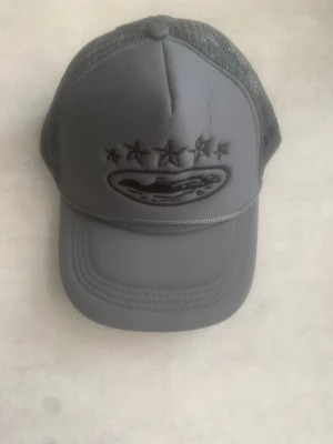 Grå meshkeps med broderade stjärnor - Snygg grå keps med mesh på sidorna och bak, samt broderade svarta stjärnor och ett ovalt märke framtill. Böjd skärm och justerbar passform. Perfekt för en avslappnad streetstyle-look.