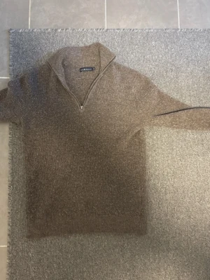 Beige stickad tröja med dragkedja - Stickad tröja från GANT i beige nyans med halv dragkedja framtill och hög krage. Tröjan har ribbstickad struktur och långa ärmar, perfekt för lager-på-lager-stil. Klassisk och stilren design som funkar till mycket.