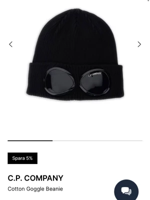 Svart Goggle Beanie från C.P. Company -  svart mössa från C.P. Company med deras ikoniska goggle-detaljer på uppviket. Mössan är ribbstickad och tillverkad i bomull, vilket gör den både bekväm och stilren. Nypris 1200kr