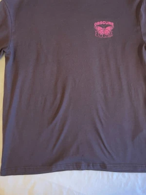 Lila t-shirt Obscure Future fjäril - Lila t-shirt från Obscure Future med tryck av en rosa fjäril och texten 'Fly like a butterfly' på bröstet. T-shirten har rund hals och är tillverkad i mjuk bomull. Perfekt för dig som gillar streetwear och coola prints.