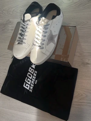 Vita Golden Goose sneakers med stjärna - Snygga vita sneakers från Golden Goose med grå mockastjärna på sidan, grå skosnören och svart hälparti. Skorna har en klassisk låg modell och är tillverkade i läder med detaljer i mocka. Perfekta för dig som gillar en trendig och avslappnad stil.