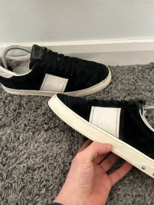 Svarta och vita sneakers från Valentino - Snygga sneakers från Valentino i svart mocka med vita detaljer och vit sula. Klassisk låg modell med svarta skosnören och diskret logga på plösen. Perfekta för dig som vill ha en stilren och exklusiv look. Lite slitna på bakdelen inne i skon som syns på bilden och det är så på båda sidorna men de finns en lapp där som täcker vilket gör att de inte påverkar så mycket, annars bra skick kommer med dustbag