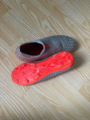 Gråa Puma Netfit fotbollsskor - Säljer ett par gråa Puma Netfit fotbollsskor med orange detaljer och sula. Skorna har en stickad ovandel med struktur för bättre bollkontroll och en låg strumpliknande passform. Perfekta för dig som vill sticka ut på planen med både stil och teknik.