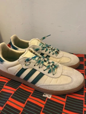 Adidas Wales Bonner Samba sneakers - Säljer ett par Adidas Wales Bonner Samba sneakers i vitt och beige med gröna detaljer och klassiska tre ränder på sidan. Skorna har mönstrade gröna/vita skosnören och gummisula. Materialmix av mocka och läder ger en retrovibe. Perfekta för dig som gillar unik streetstyle.