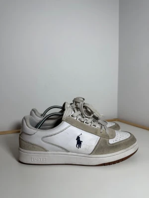 Vita och beige sneakers från Polo Ralph Lauren - Snygga sneakers i vitt och beige från Polo Ralph Lauren med klassisk logga på sidan. Skorna har ovandel i läder och mocka, perforerad tå och vita snören. Yttersulan är vit med brun undersida. Perfekt för dig som gillar stilrena och sportiga sneakers.