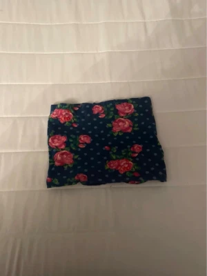 Blommig blå tubscarf - Säljer en blå tubscarf med rosa rosor och små vita prickar. Mjuk och tunn, perfekt att ha runt halsen för en färgglad touch. Mönstret är livligt och ger en retrovibe. Passar dig som vill ha något unikt och lekfullt i din stil.