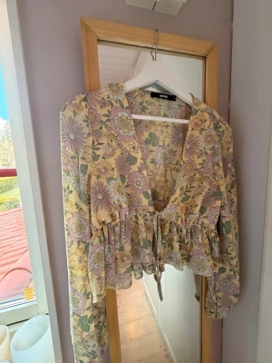 Blommig blus med volang och knyt - Supersöt blommig blus från Bershka i pastelliga toner. Blusen har v-ringning, volangdetalj nedtill och knyt framtill. Långärmad och croppad modell, perfekt för vår och sommar.