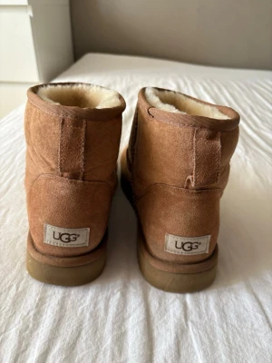 Bruna UGG Classic Mini boots - Mysiga bruna UGG Classic Mini boots i mocka med mjukt vitt fårskinnsfoder och grov gummisula. Perfekta för kalla dagar och ger en avslappnad och trendig look. Klassisk UGG-logga baktill och låg skaftmodell.