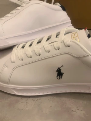 Vita sneakers från Polo Ralph Lauren - Säljer vita sneakers från Polo Ralph Lauren med klassisk logga i svart på sidan. Skorna har vita snören, detaljer i guld och mörkblått samt röd och blå rand på hälen. Köpte från när jag var på semester i frankrike och säljer de för ett billigare pris än originalet, och har aldrig använt de.