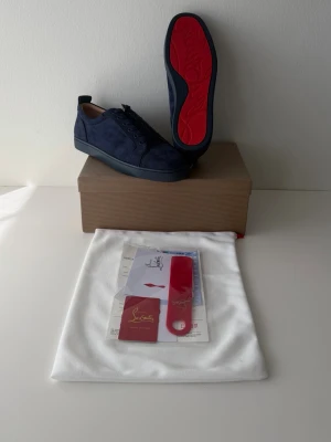 Blåa sneakers från Christian Louboutin - Snygga mörkblå sneakers från Christian Louboutin i mocka med den ikoniska röda sulan. Skorna har klassisk låg profil, rund tå och matchande blå snörning. Kommer med originalpåse, skohorn och certifikat. Perfekta för dig som vill sticka ut med exklusiv stil.