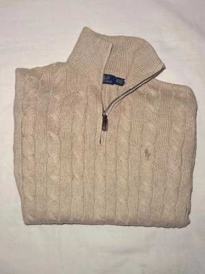 Beige kabelstickad tröja Polo Ralph Lauren - Kabelstickad beige tröja från Polo Ralph Lauren med halv dragkedja och hög krage. Tröjan har ribbade muddar vid ärmslut och nederkant samt den klassiska broderade loggan på bröstet. Perfekt för lager på lager och riktigt snygg till jeans.