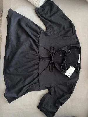 Svart peplum blus med knytning från lager 157 - Stilren svart blus från 157 med knytning framtill och lätt puffade trekvartsärmar. Blusen har en lös passform och är tillverkad i ett mjukt, lätt glansigt tyg som ger en snygg siluett. Perfekt att matcha med jeans eller kjol för en trendig look. Blusen är helt ny med prislapp, oanvänd.